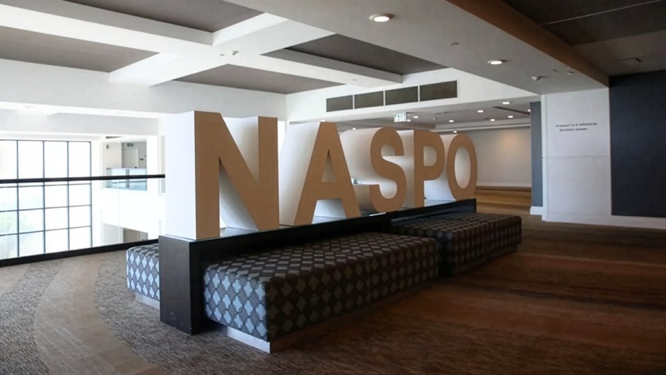 Large freestanding NASPO letters sit atop patterned bench seating in a spacious lobby area. The setting features a modern atrium with high ceilings, recessed lighting, and wide windows providing natural light. Neutral tones of brown, gray, and white create a clean, professional atmosphere. The space appears designed for conference attendees or visitors to gather and relax.
