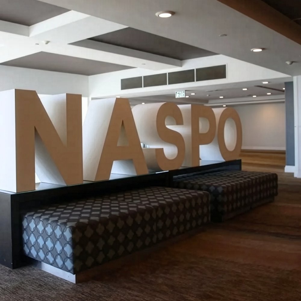 Large freestanding NASPO letters sit atop patterned bench seating in a spacious lobby area. The setting features a modern atrium with high ceilings, recessed lighting, and wide windows providing natural light. Neutral tones of brown, gray, and white create a clean, professional atmosphere. The space appears designed for conference attendees or visitors to gather and relax.