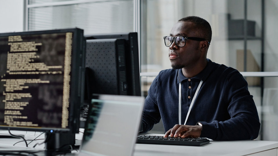 African American young developer in eyeglasses concentrating on his online work on computer sitting at workplace