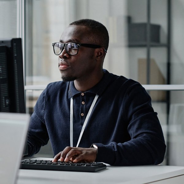 African American young developer in eyeglasses concentrating on his online work on computer sitting at workplace