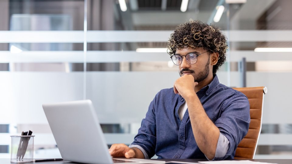 A thoughtful young Indian man is sitting at a desk in the office and looking at the laptop screen.