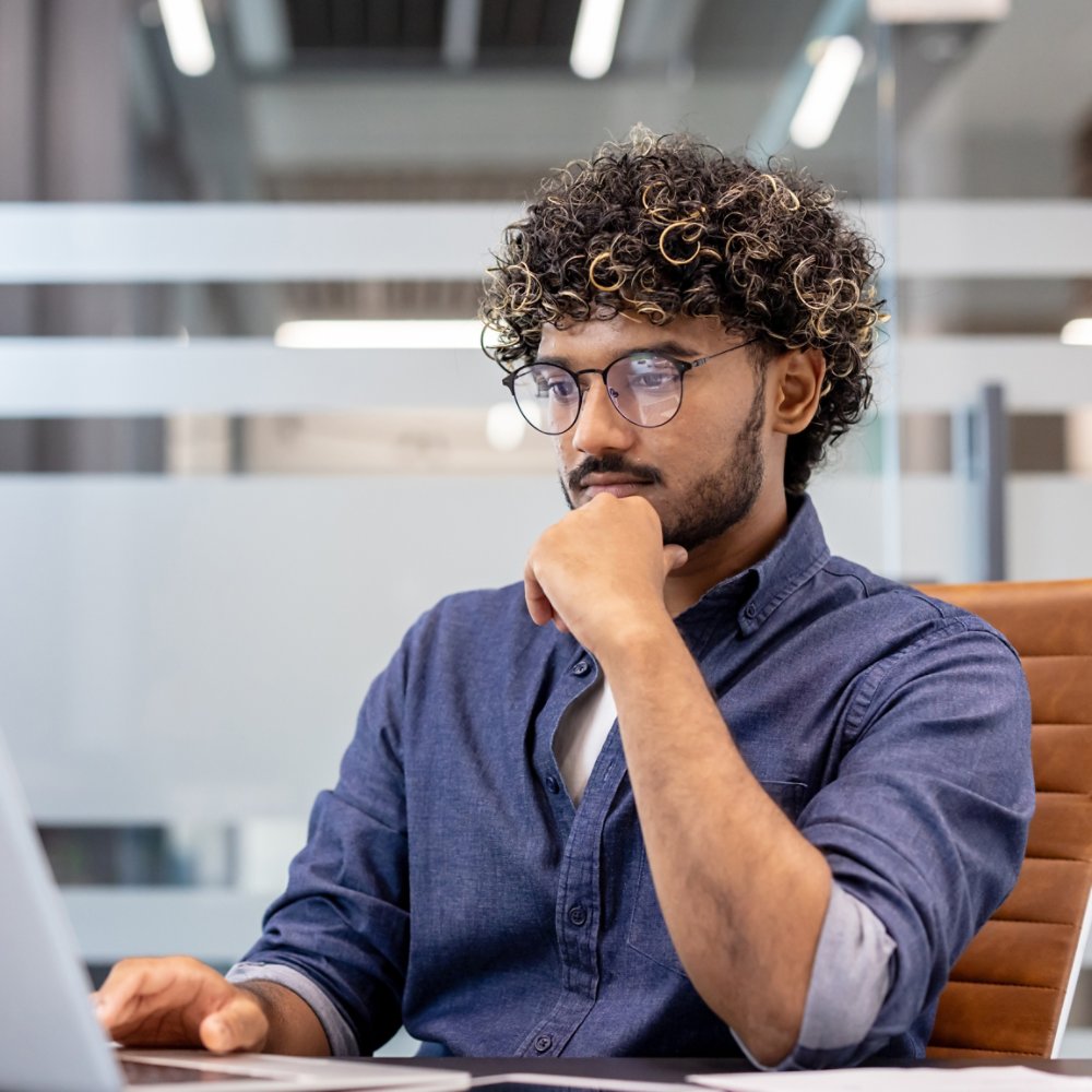 A thoughtful young Indian man is sitting at a desk in the office and looking at the laptop screen.