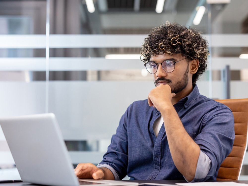 A thoughtful young Indian man is sitting at a desk in the office and looking at the laptop screen.