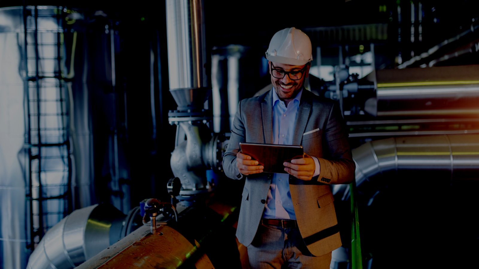 Handsome smiling caucasian supervisor in suit and with helmet on head using tablet while standing in power plant.