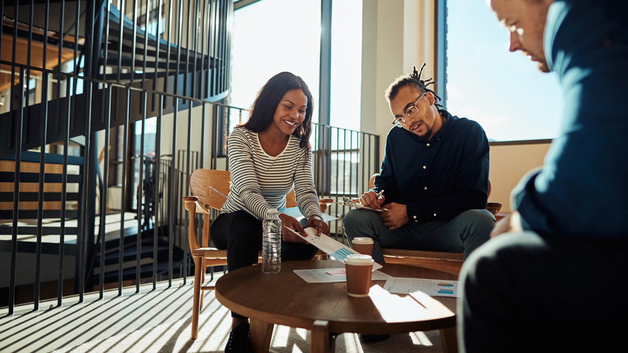 Two smiling young African American businesspeople discussing paperwork with colleagues during a meeting in the lounge of a modern office