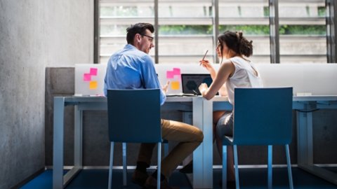 An attractive woman indoors talking to a handsome man sitting at the desk, discussing an issue.