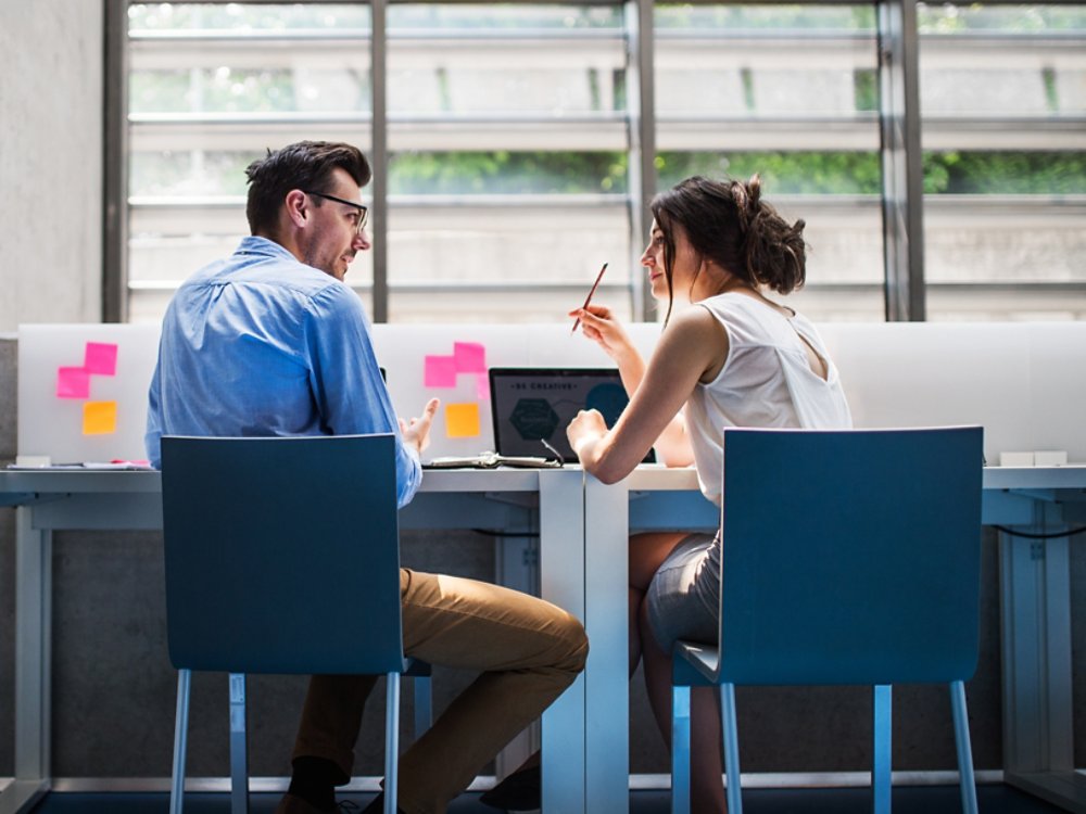 An attractive woman indoors talking to a handsome man sitting at the desk, discussing an issue.