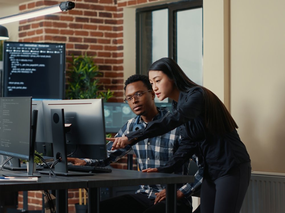 Two software developers holding laptop with coding interface walking towards desk and sitting down talking about group project. Programmers team discussing algorithms pointing at computer screen.