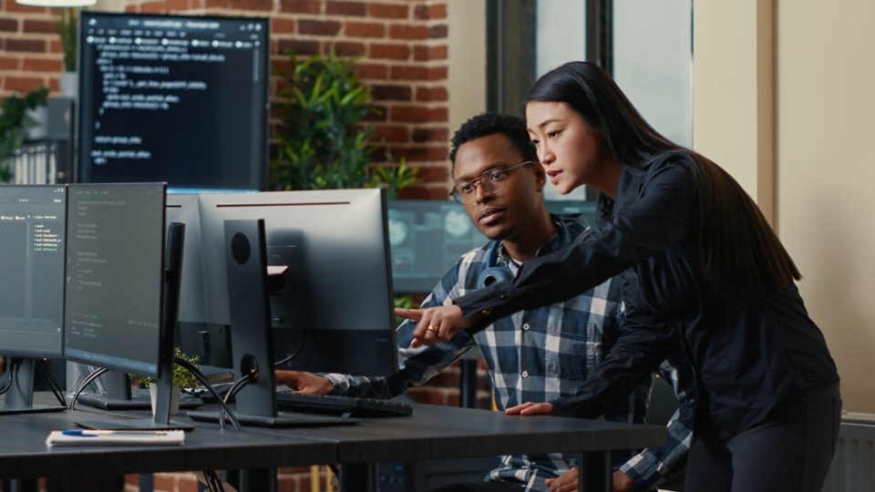 Two software developers holding laptop with coding interface walking towards desk and sitting down talking about group project. Programmers team discussing algorithms pointing at computer screen.