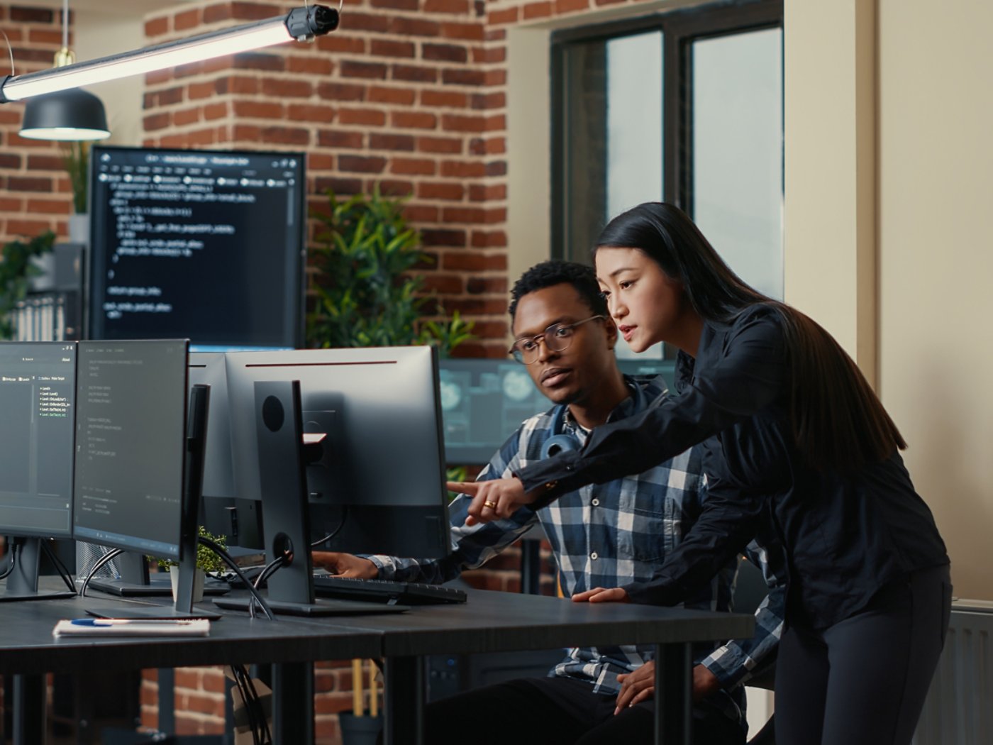 Two software developers holding laptop with coding interface walking towards desk and sitting down talking about group project. Programmers team discussing algorithms pointing at computer screen.