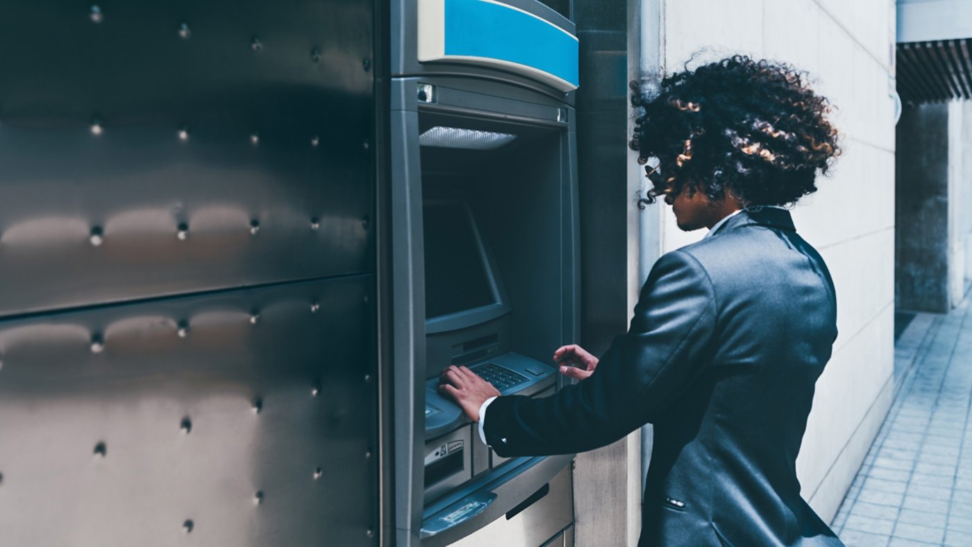 Side view of a businessman withdrawing money from his bank card using an outdoor ATM;  a curly man entrepreneur is using automated teller machine on the street to to replenish his account deposit