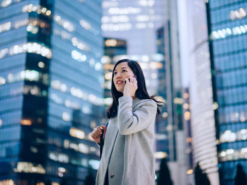 Confident and successful young Asian businesswoman talking on smartphone with business partners on the go, against contemporary corporate skyscrapers with illuminated facade in financial district in the evening. Business on the go