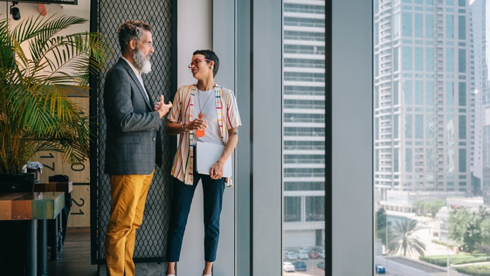Two colleagues stand and talk near large floor-to-ceiling windows in a modern office. One person in a blazer and yellow pants gestures while speaking, and the other holds a tablet and listens, smiling. Tall city skyscrapers and palm trees are visible outside, suggesting an urban business district. Indoor plants and contemporary interior details add a relaxed, creative atmosphere.
