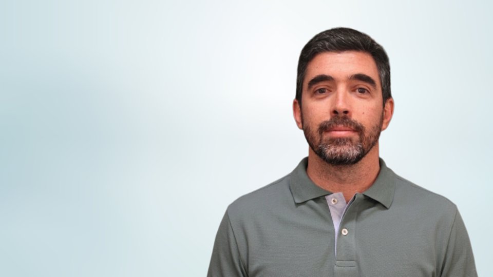 A man with short dark hair and a trimmed beard faces the camera in a straightforward portrait. He is wearing a gray polo shirt with a light-colored placket and buttons. The background is a soft, light gradient that keeps the focus on the subject. The overall composition is simple and professional, suitable for profile or corporate use.