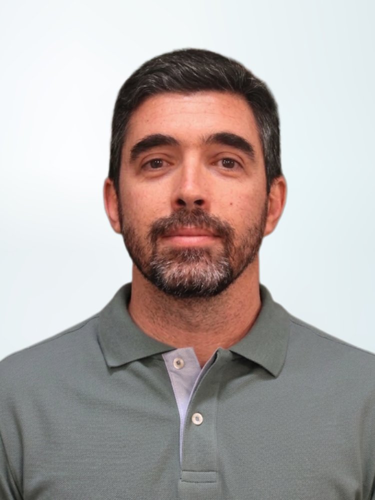 A man with short dark hair and a trimmed beard faces the camera in a straightforward portrait. He is wearing a gray polo shirt with a light-colored placket and buttons. The background is a soft, light gradient that keeps the focus on the subject. The overall composition is simple and professional, suitable for profile or corporate use.