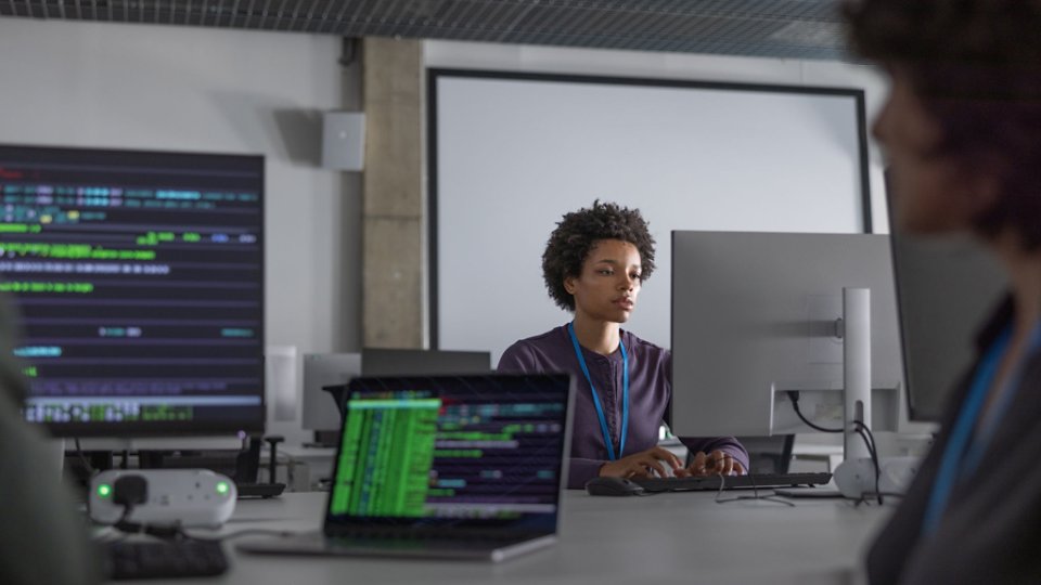 A person sits at a desk in a modern office, concentrating on a large desktop monitor. Multiple screens around the room display colorful code and data, suggesting a software development or cybersecurity environment. The setting appears collaborative, with other colleagues partially visible in the foreground. Cool, muted lighting and neutral tones create a professional, tech-focused atmosphere.