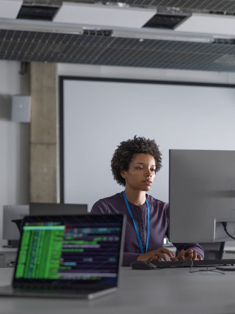 A person sits at a desk in a modern office, concentrating on a large desktop monitor. Multiple screens around the room display colorful code and data, suggesting a software development or cybersecurity environment. The setting appears collaborative, with other colleagues partially visible in the foreground. Cool, muted lighting and neutral tones create a professional, tech-focused atmosphere.