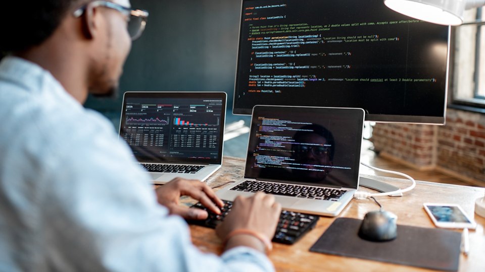 Young african male programmer writing program code sitting at the workplace with three monitors in the office. Image focused on the screen