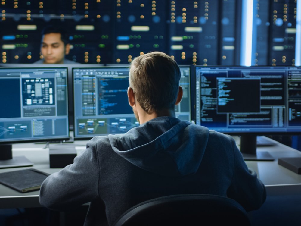 IT Specialist Works on Personal Computer with Screens Showing Software Program with Coding Language Interface. In the Background Technical Room of Data Center with Professional Working