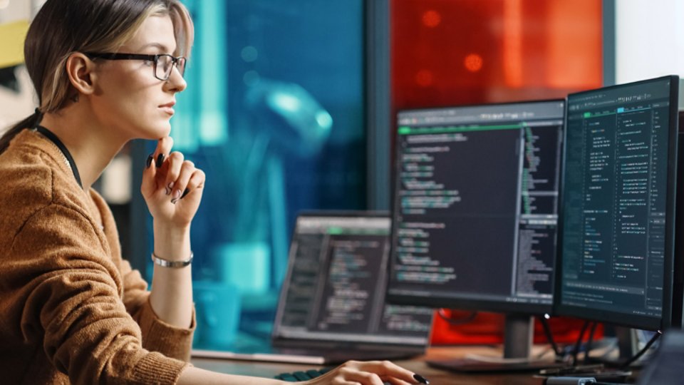 Female Junior Software Engineer Writes Code on Desktop Computer With Two Monitors and Laptop Aside In Stylish Office. Caucasian Woman Working On Artificial Intelligence Service For Big Tech Company.