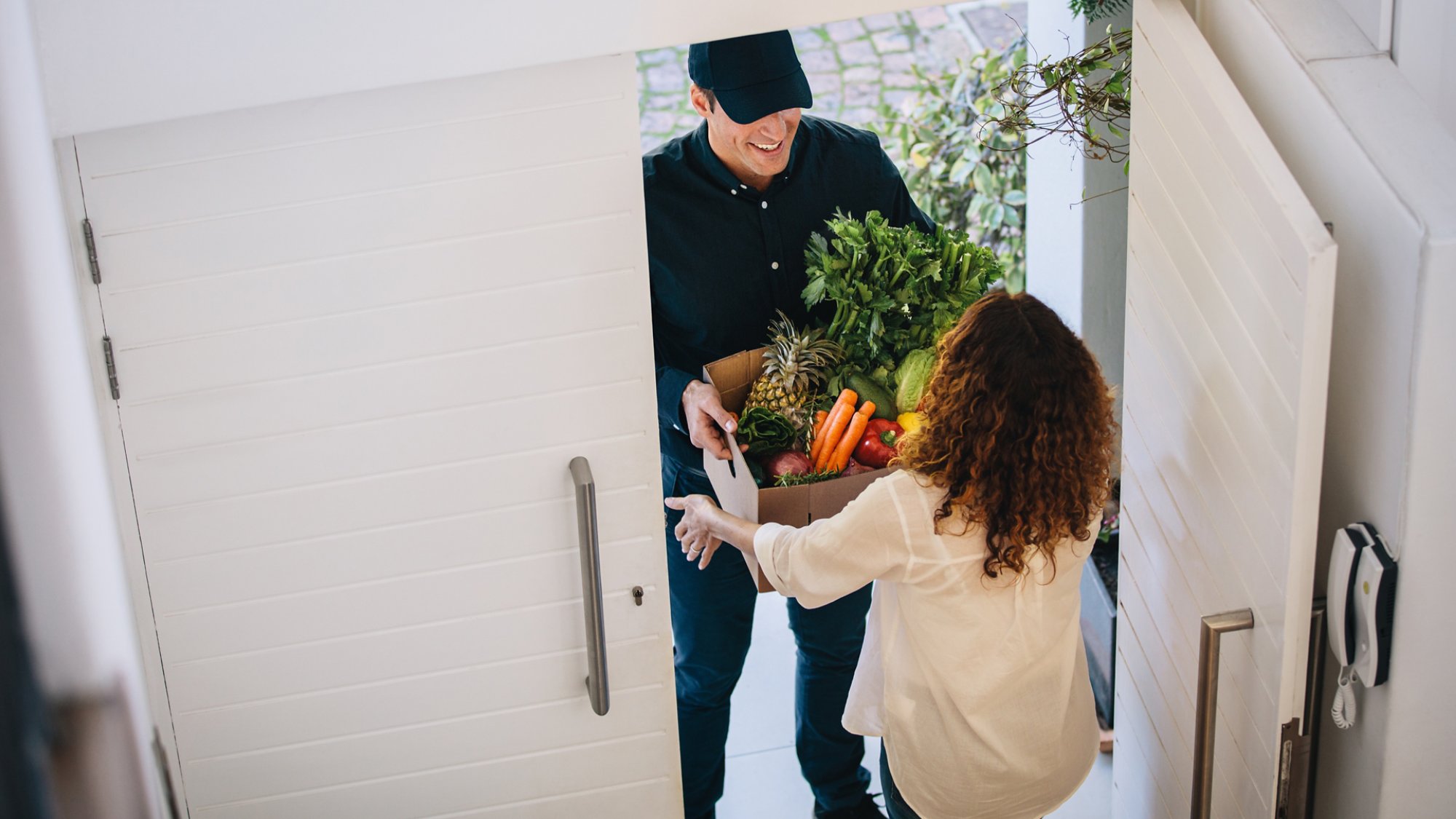 High angle view of a female customer receiving groceries from delivery man at the house's door. Delivery employee delivering a box of fruits and vegetables to a woman.