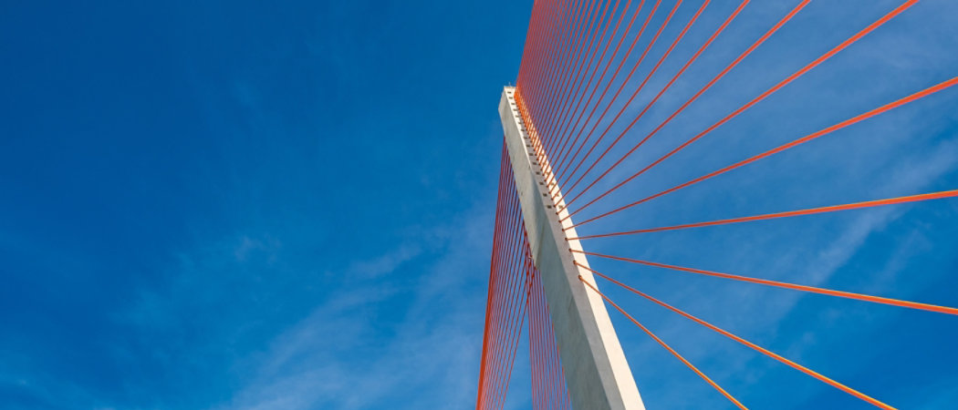 A striking cable-stayed bridge with orange suspension cables extending from a tall concrete pillar. The structure is set against a vibrant blue sky with scattered clouds, emphasizing its modern architectural design. The upward perspective highlights the geometric lines and engineering precision.