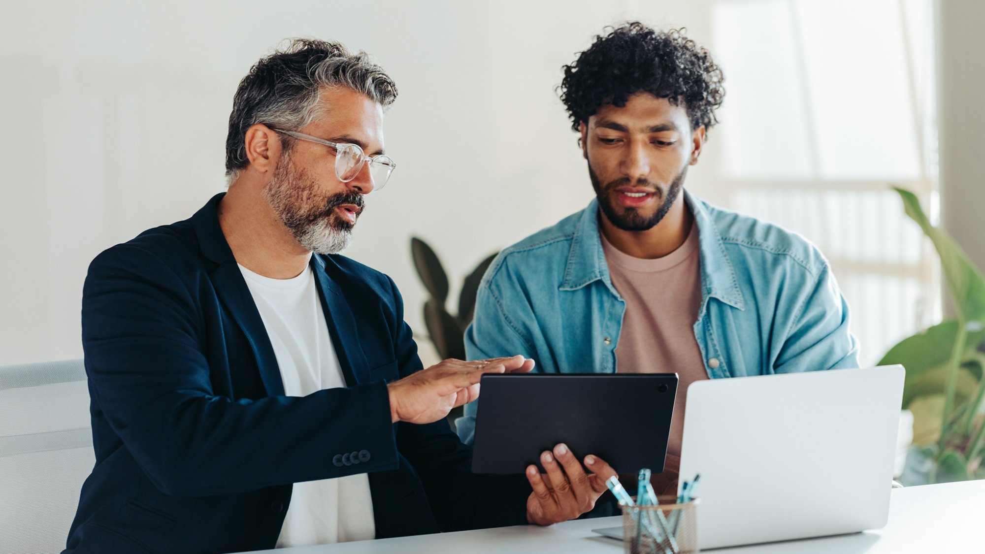 Professional men are engaged in a productive discussion in a brightly lit office. They use a tablet and a laptop to discuss and brainstorm business matters, highlighting teamwork and collaboration.