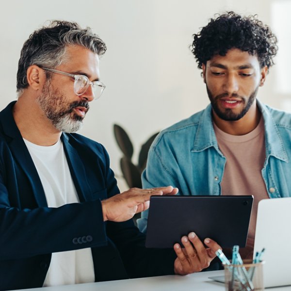 Professional men are engaged in a productive discussion in a brightly lit office. They use a tablet and a laptop to discuss and brainstorm business matters, highlighting teamwork and collaboration.