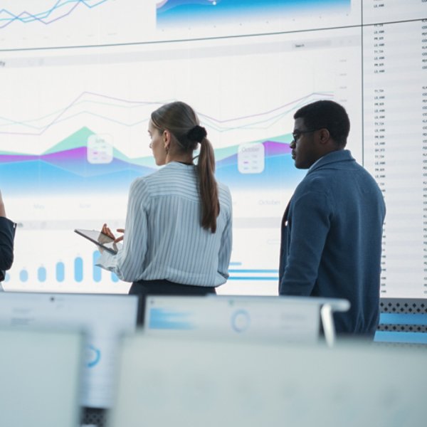 Diverse Team Of Data Scientists Having A Meeting And Discussing Reports In a Modern Monitoring Office With Analytics Feed On a Big Digital Screen. Deep Learning Company With Employees Behind Computers