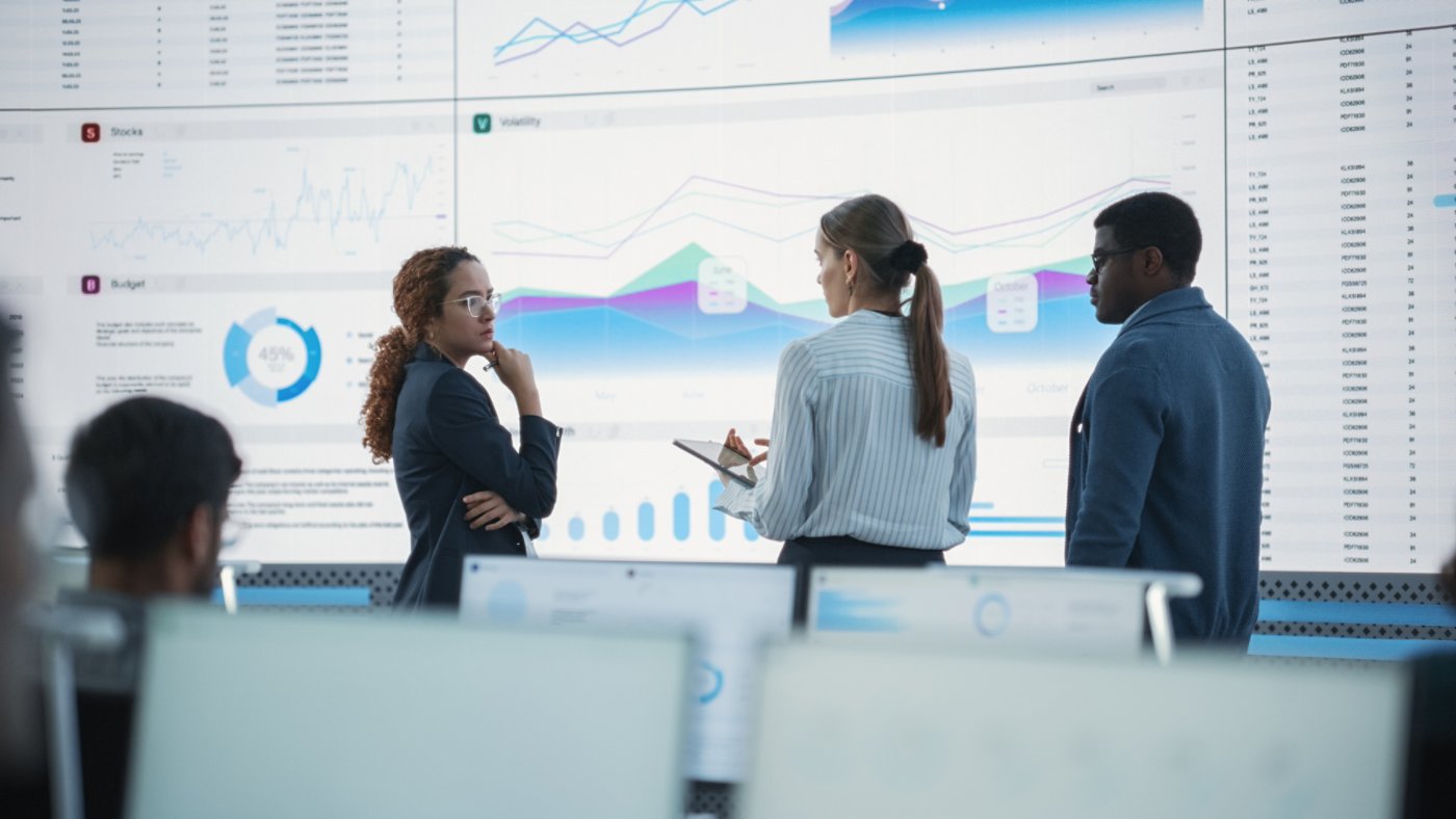 Diverse Team Of Data Scientists Having A Meeting And Discussing Reports In a Modern Monitoring Office With Analytics Feed On a Big Digital Screen. Deep Learning Company With Employees Behind Computers