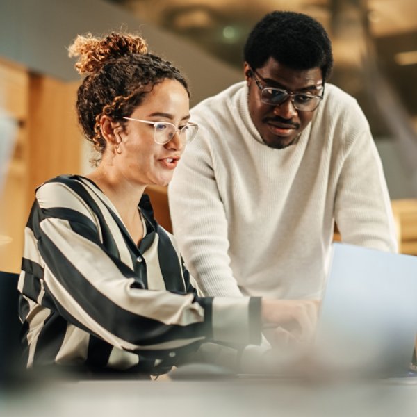 Portrait of Two Creative Colleagues Using Laptop to Discuss Work Project at Office. Young Black Technical Support Specialist Helping Female Customer Relationship Coordinator. Teamwork Concept