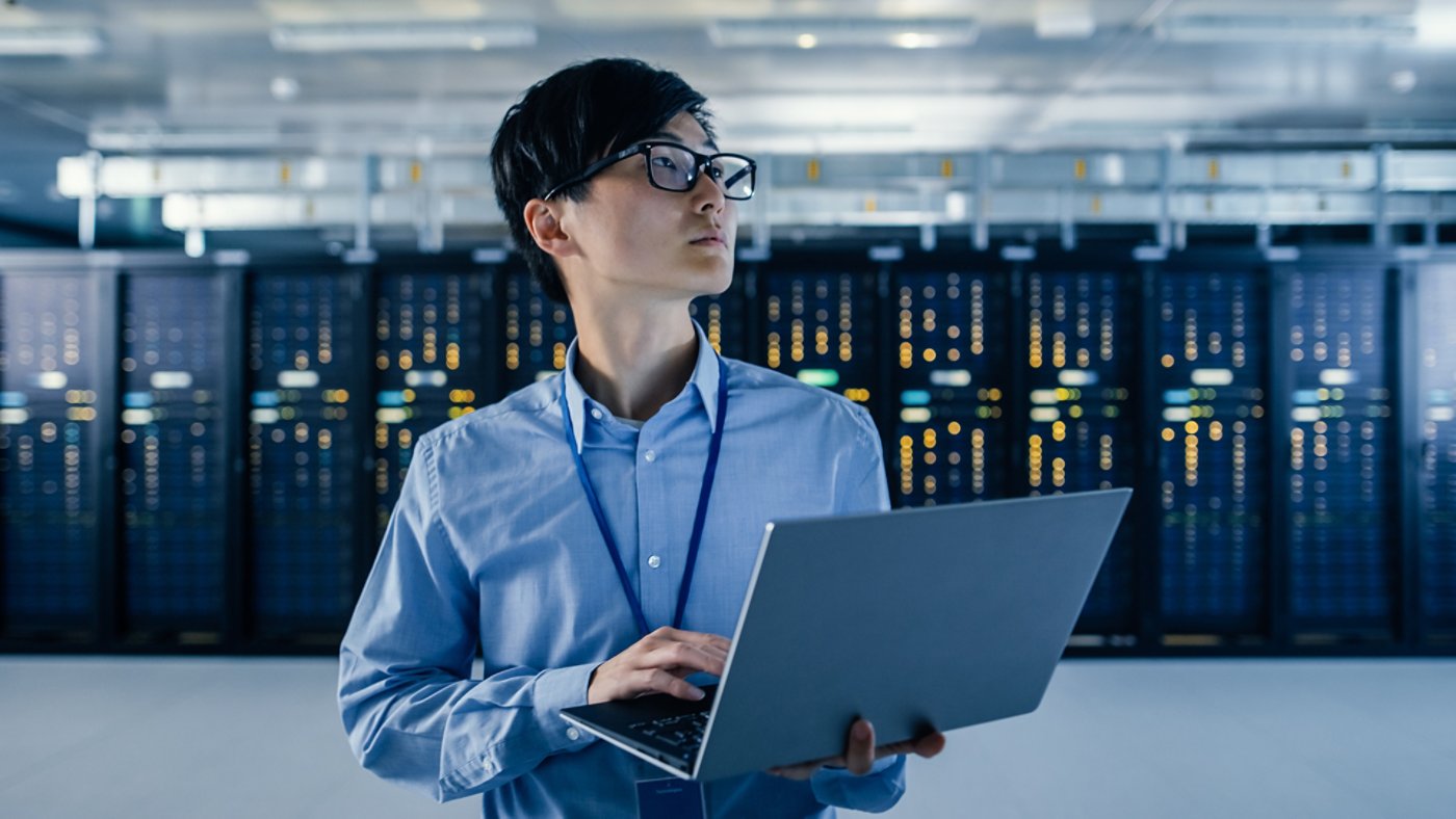 In the Modern Data Center: Portrait of IT Engineer Stand Near Server Racks, Finishing Maintenance and Diagnostics Procedure Holding Laptop.