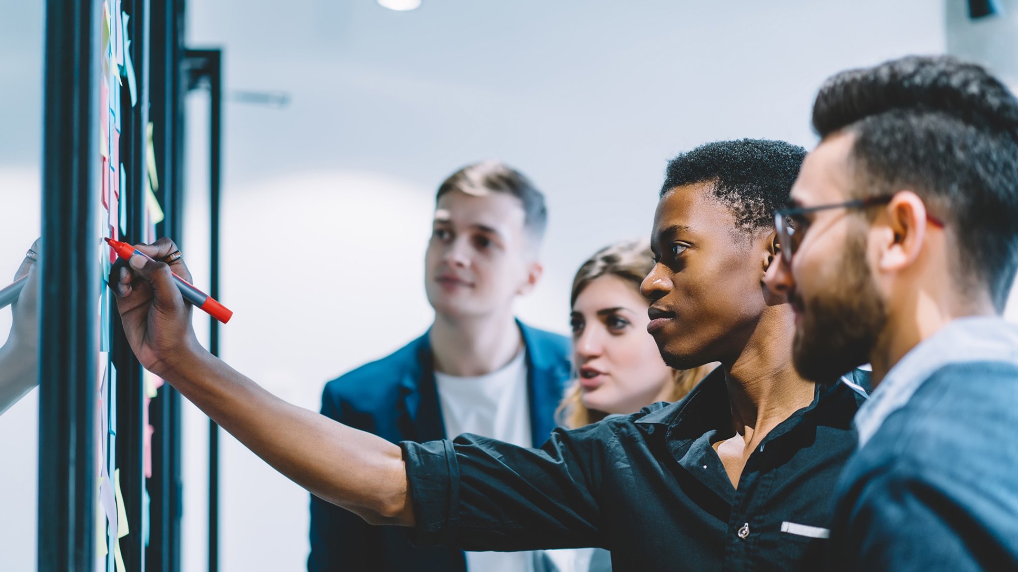 Diverse team of skilled students together solving task writing down information on papers glued on wall.Multicultural young people collaborating on business plan making notes of ideas on sticker