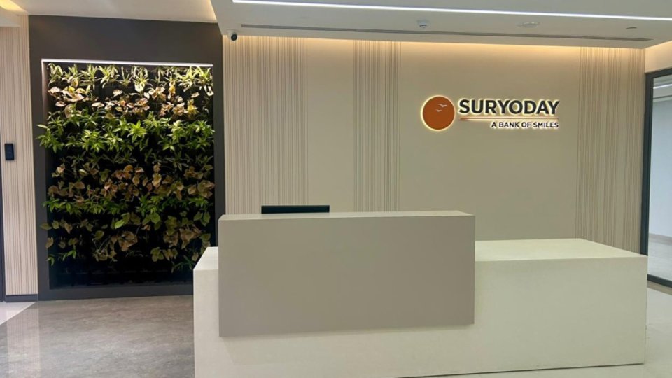 A contemporary reception desk stands in a bright office lobby. The wall behind the desk features the Suryoday name and tagline "A Bank of Smiles" beside an orange circular emblem. Vertical textured panels add subtle design detail to the light-colored interior. On the left, a lush vertical garden brings greenery and contrast to the clean, minimalist space.