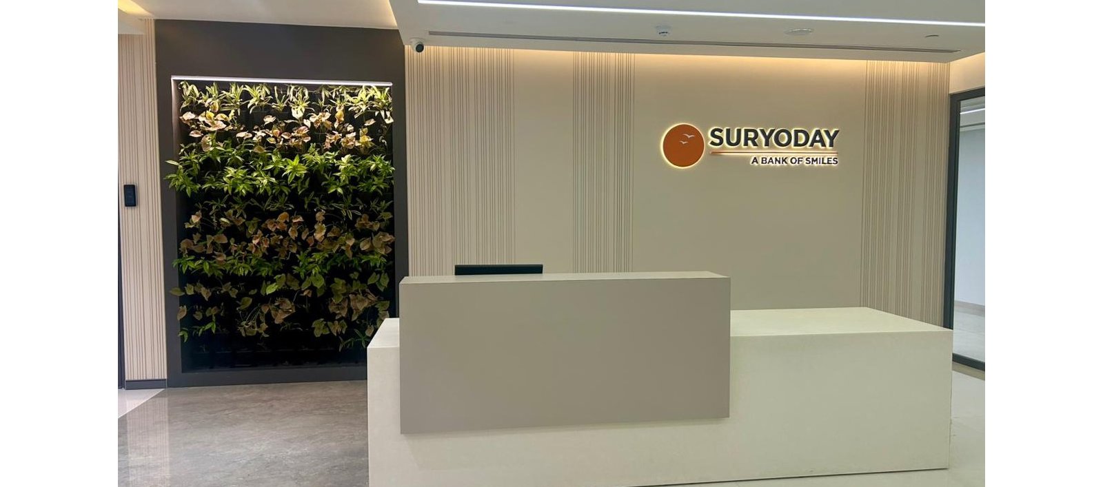 A contemporary reception desk stands in a bright office lobby. The wall behind the desk features the Suryoday name and tagline "A Bank of Smiles" beside an orange circular emblem. Vertical textured panels add subtle design detail to the light-colored interior. On the left, a lush vertical garden brings greenery and contrast to the clean, minimalist space.