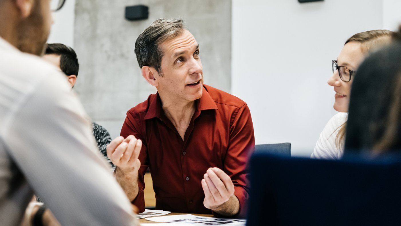 A caucasian man is speaking in a business meeting surrounded by colleagues listening to him. He uses his hands and seems convinced and focused.