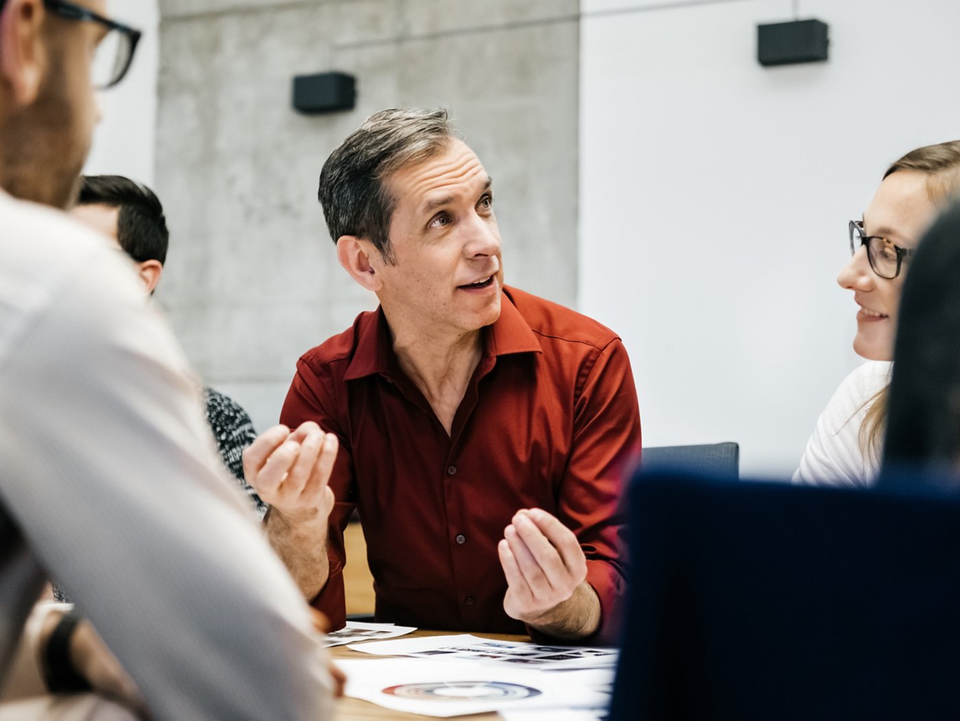 A caucasian man is speaking in a business meeting surrounded by colleagues listening to him. He uses his hands and seems convinced and focused.