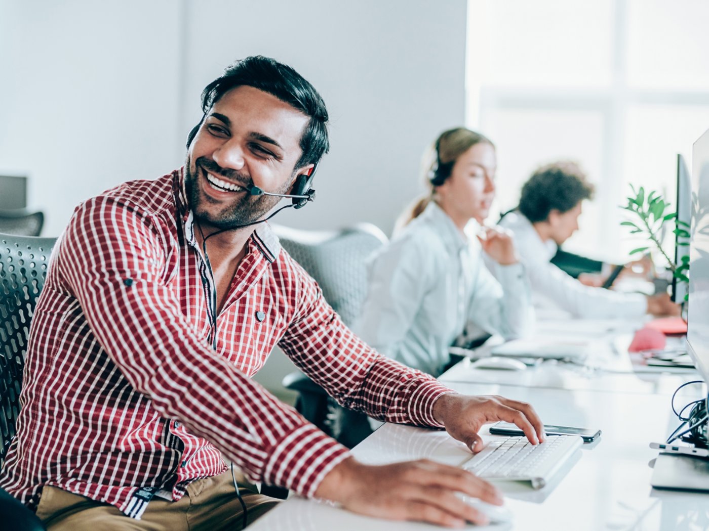 Smiling handsome ethnicity businessman working in call center. Shot of a cheerful young man working in a call center with his team. Confident male operator is working with colleagues. Call center operators sitting in a row at desks.