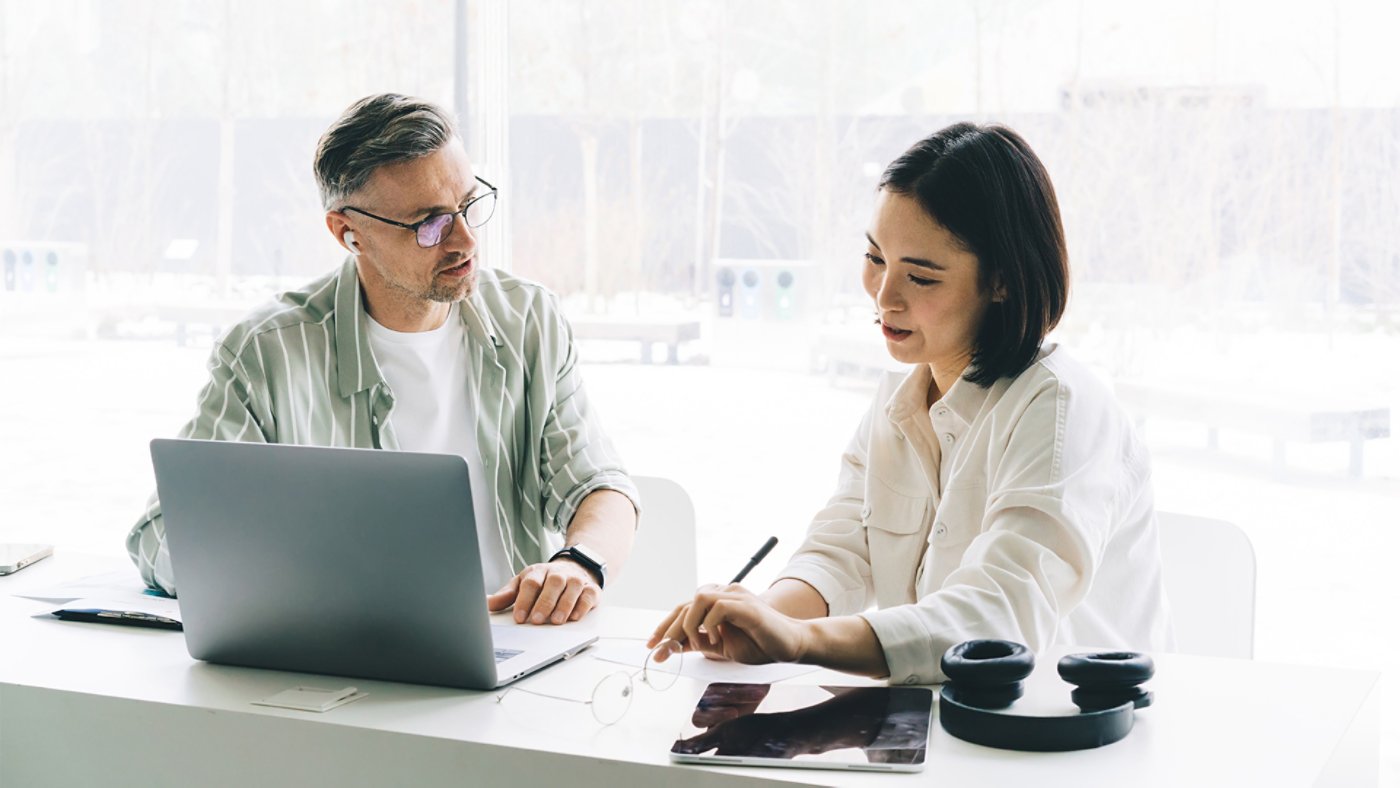 Male employee in optical eyeglasses communicate with Asian female colleague during cooperation process at table desktop with laptop technology, professional entrepreneurs talking about business