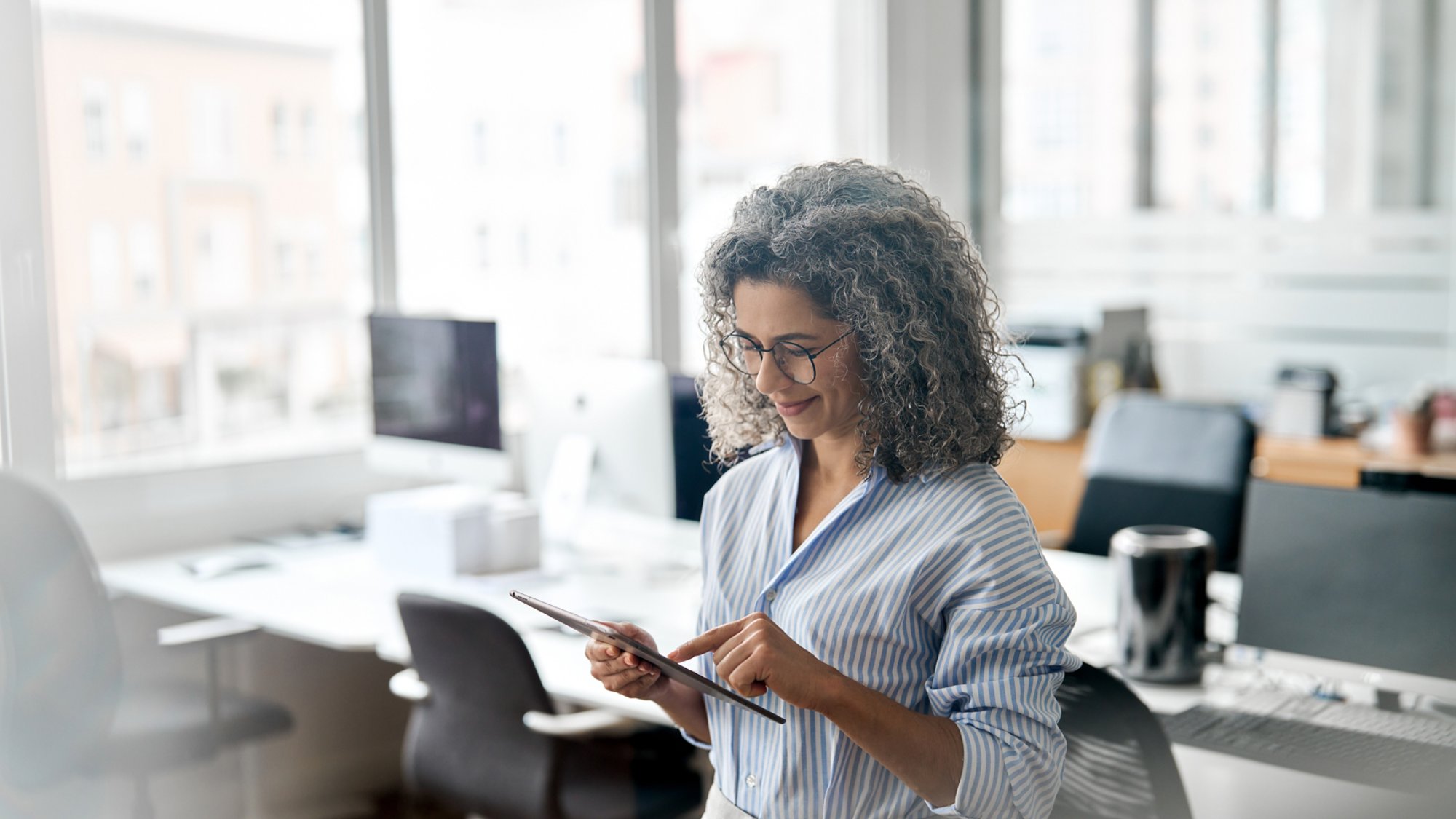 Busy middle aged professional business woman using tab computer in office. Mature lady manager, older female corporate executive holding tablet standing at work, authentic shot. View through glass