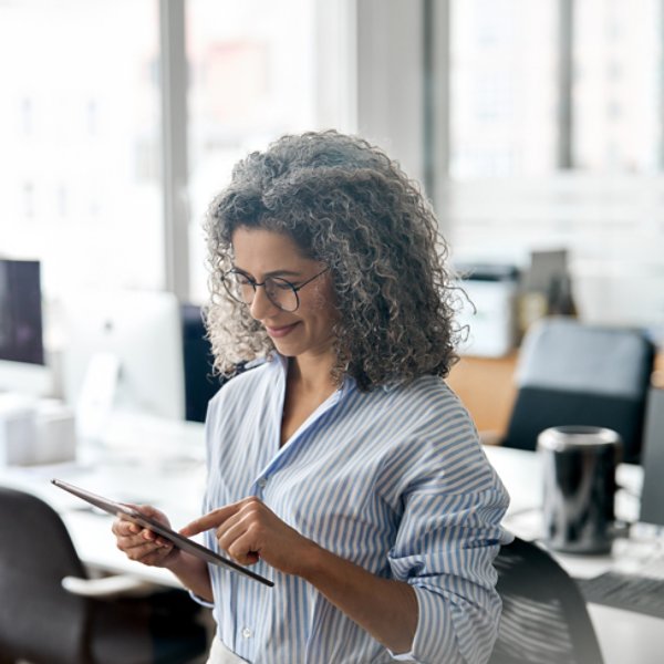 Busy middle aged professional business woman using tab computer in office. Mature lady manager, older female corporate executive holding tablet standing at work, authentic shot. View through glass