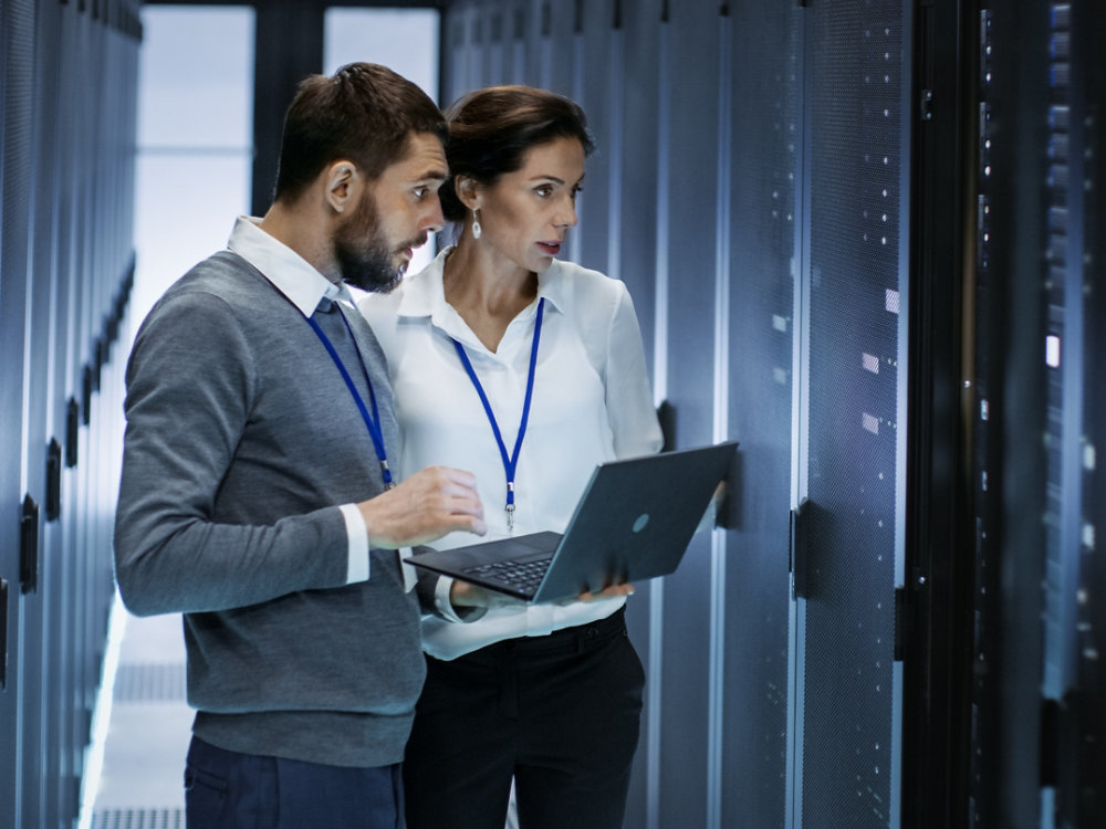 Male IT Specialist Holds Laptop and Discusses Work with Female Server Technician. They're Standing in Data Center, Rack Server Cabinet is Open.