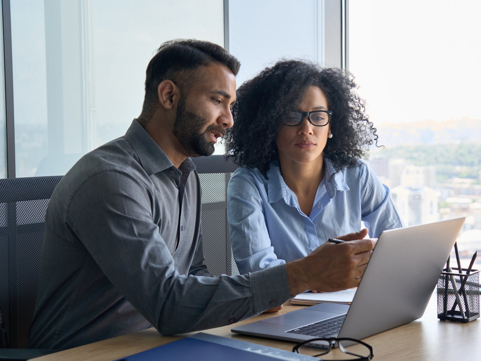 Indian male ceo executive manager mentor giving consultation on financial operations to female African American colleague intern using laptop sitting in modern office near panoramic window.