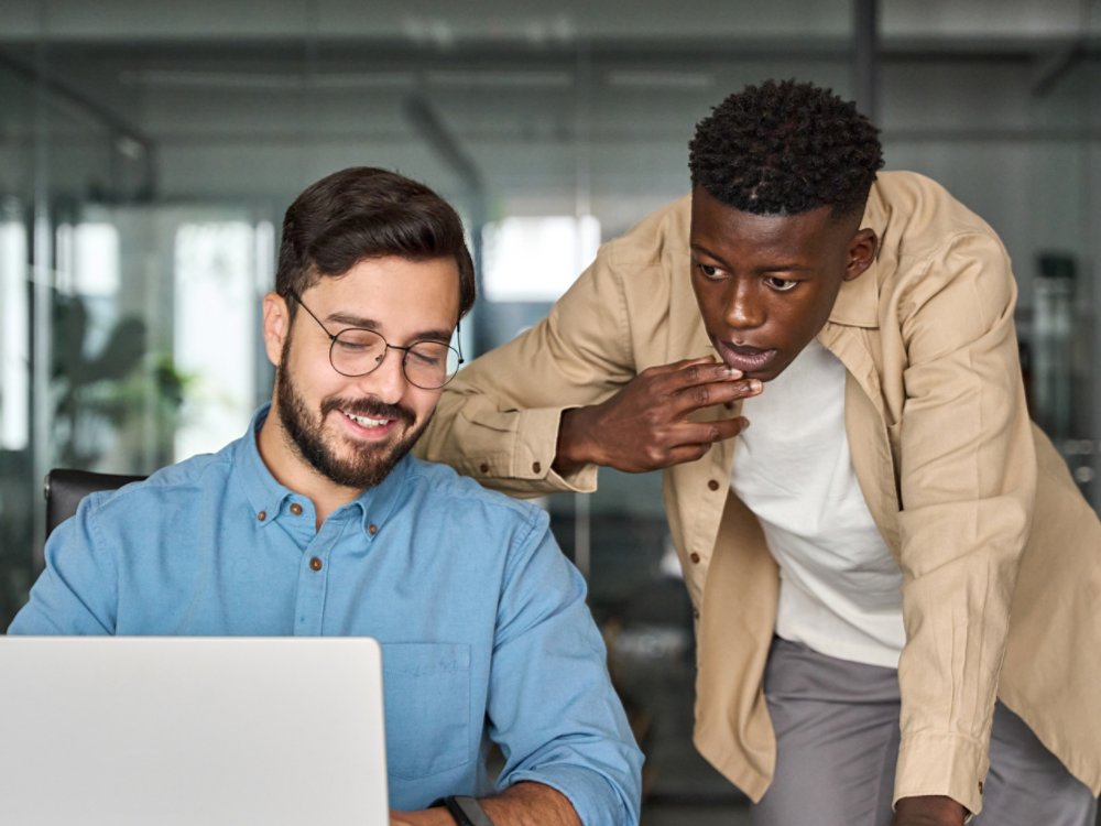 Two busy diverse professional male coworkers software engineers team discussing project together working in teamwork talking sharing ideas sitting at work desk with laptop computer.