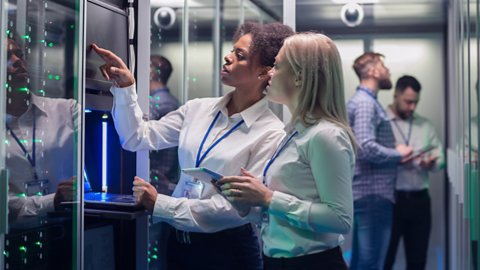Wide shot of casual man with tablet walking among server racks in data center room looking at hardware