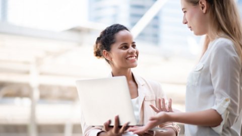 two women outdoors discussing business holding laptop-16x9