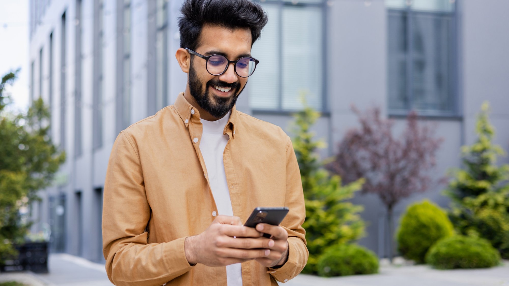 Successful Indian young businessman outside office building walking in daytime, man holding phone in hands, businessman dialing, browsing online pages, programmer engineer in glasses.