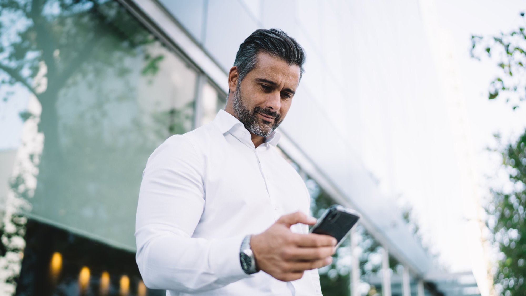 Below view of mature lawyer with stubble using mobile application for chatting in business chat, Caucasian male proud CEO in white shirt searching banking app for making online payment and banking
