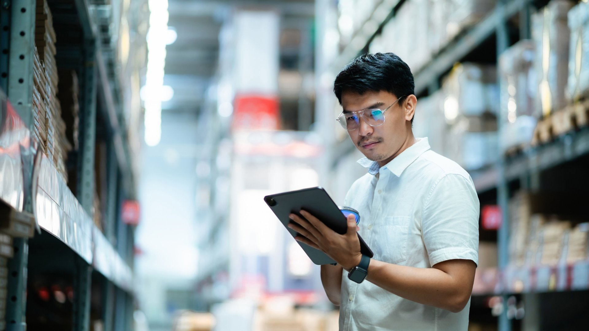 A man wearing glasses is looking at a tablet in a warehouse. He is focused on the screen and he is working