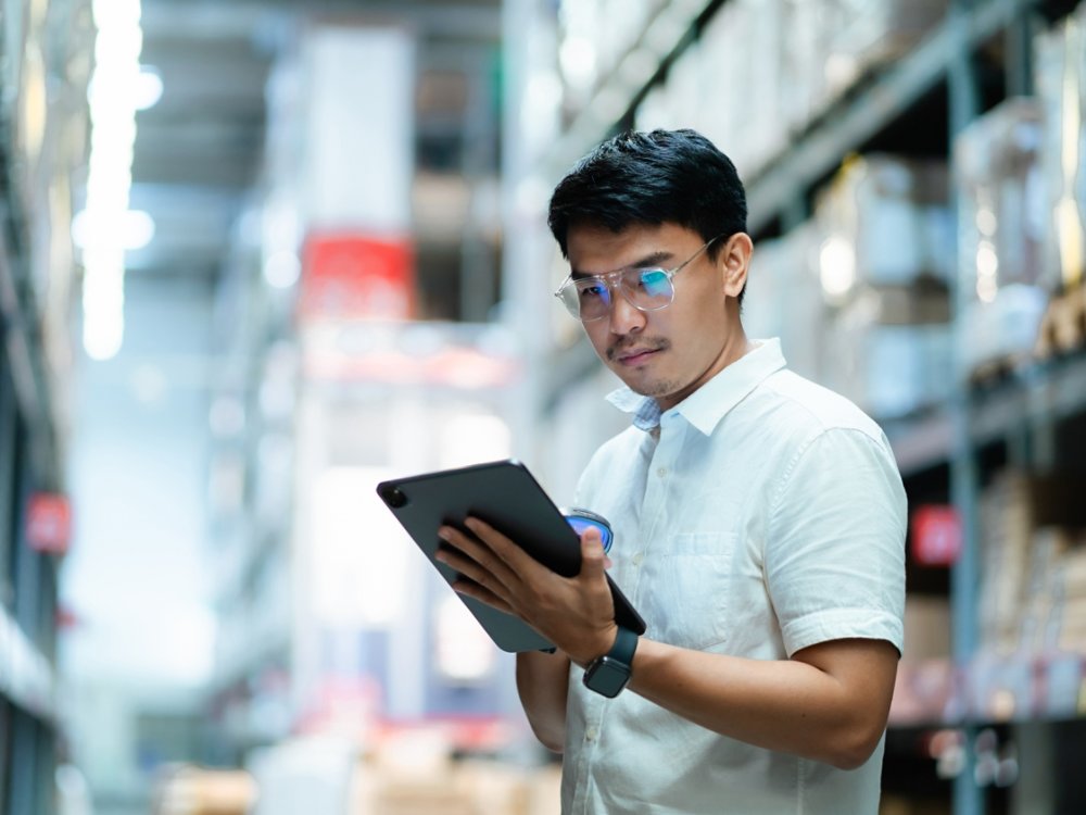 A man wearing glasses is looking at a tablet in a warehouse. He is focused on the screen and he is working